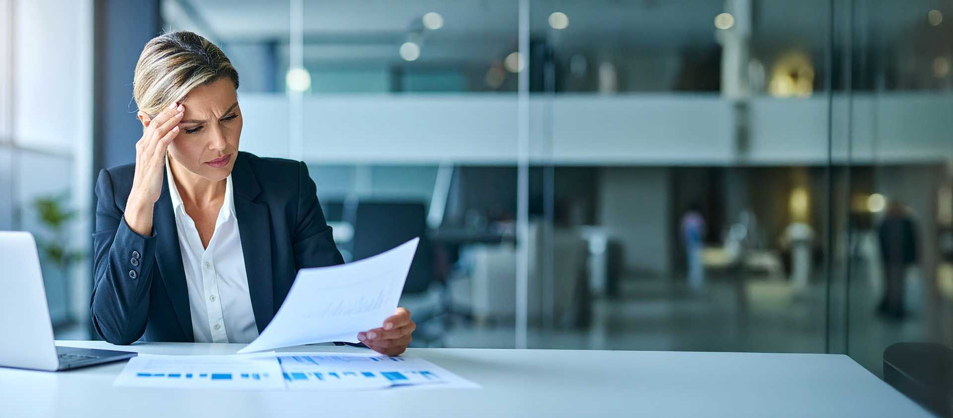 A woman in professional business attire sits at her desk, her gaze fixed with apprehension on the financial documents before her.